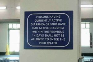 A blue and white sign on a white wall with the text "PERSONS HAVING CURRENTLY ACTIVE DIARRHEA OR WHO HAVE HAD ACTIVE DIARRHEA WITHIN THE PREVIOUS 14 DAYS SHALL NOT BE ALLOWED TO ENTER THE POOL WATER."