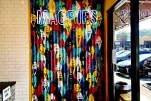 A photorealistic image of a vibrant indoor space featuring a sidewall adorned with a whimsical hand-painted mural. Scoops of ice cream in various colors - chocolate, strawberry, vanilla, pistachio, and mint chip - are depicted in a playful pattern, each resting in a waffle cone. A neon sign with black text spelling "MAGPIE'S" is mounted on the wall, casting a soft glow on the colorful scene.
