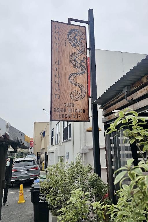 A wooden looking blade sign with the words “Pearl Dragon” against a backdrop of architectural details and a cloudy sky.