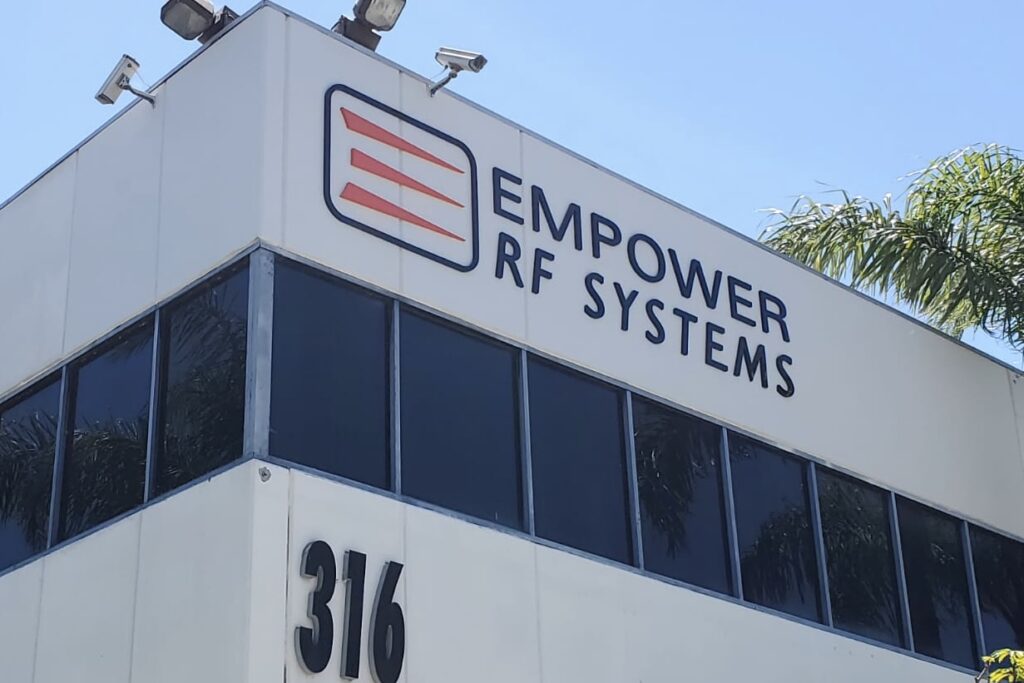 A close-up photo of a large dimensional letter sign mounted on a brick wall. The sign features bold, painted acrylic letters spelling out "Empower RF Systems" with the company logo prominently displayed in the center.