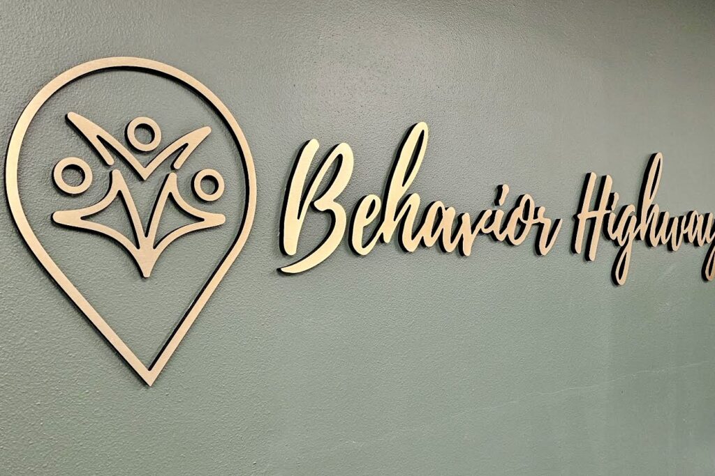 A close-up photo of the Behavior Highway lobby sign. The image focuses on the dimensional quality of the brushed gold laminate lettering and the intricate details of the company logo.