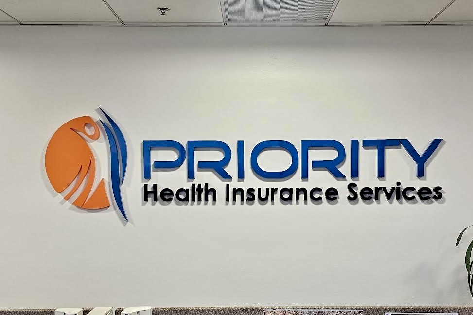 A photo of the Priority Health lobby sign mounted on a wall. The photo showcases the entire sign, highlighting the painted acrylic material, bold lettering, and prominent display of the company name and logo.