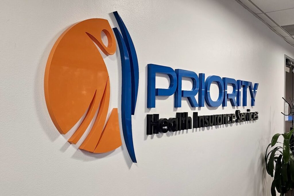 A close-up photo of the Priority Health lobby sign. The image focuses on the smooth finish of the painted acrylic and the crisp details of the lettering and logo.