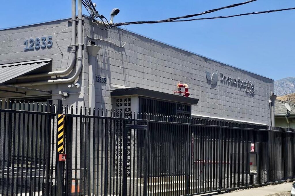 A wide shot of the Deveraux Specialties storefront. A large stainless steel sign dominates the facade, featuring the company name "Deveraux Specialties" in bold lettering with a brushed finish. The company logo sits prominently next to the  widename