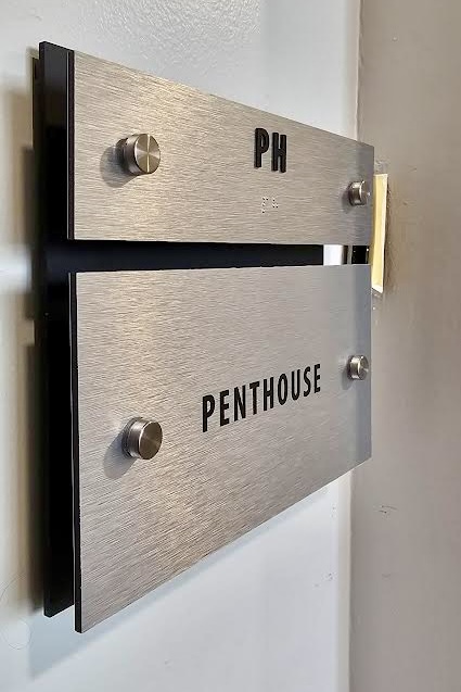 A close-up view of a penthouse suite plaque. The plaque features a brushed dibond panel with a black back panel. A tactile unit number and the tenant name are engraved on the plaque, which is mounted on the wall using standoffs.
