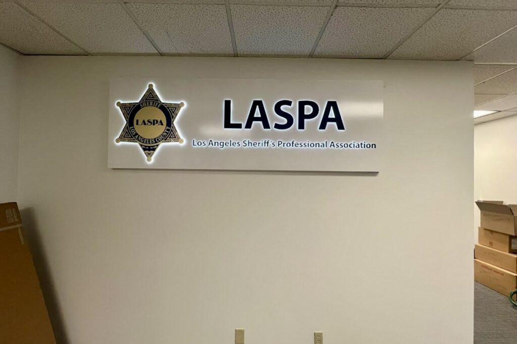 A wide shot of the LASPA lobby entrance. A custom-designed lobby sign dominates the wall behind the reception desk. The illuminated sign features the LASPA logo prominently displayed, with lettering detailing the full name "Los Angeles Sheriff's Professional Association" positioned beneath.