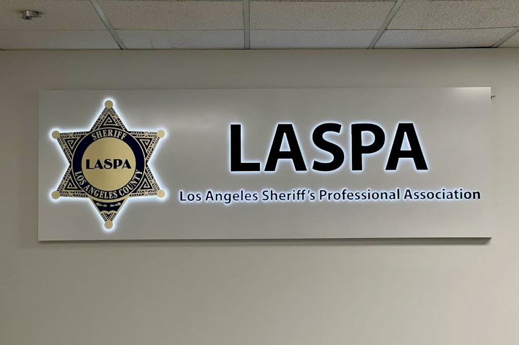 A closer-up of the LASPA lobby entrance. The wall behind the reception desk is dominated by a stunning, custom-designed lobby sign. It's illuminated and showcases the LASPA logo in a prominent display, with lettering that spells out the full name "Los Angeles Sheriff's Professional Association" positioned right beneath it. Exciting, right?