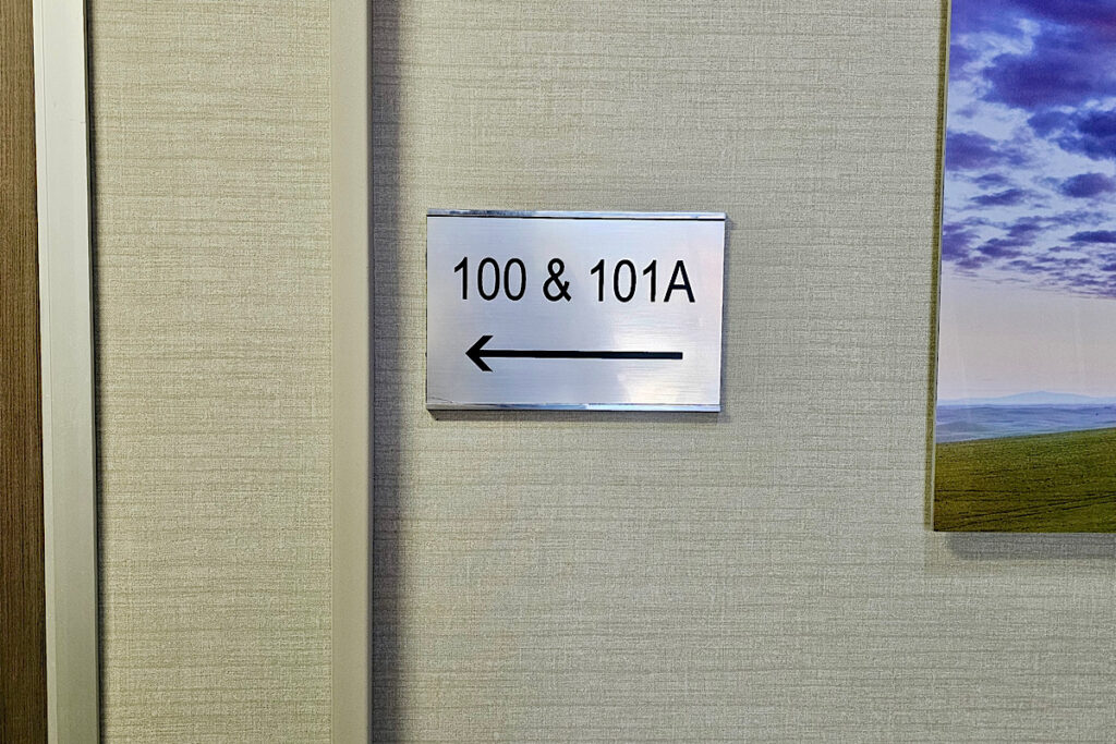 A wide shot of a hallway in the Welltower Westlake Village facility. The suite plaque inserts are mounted on the wall at eye level, clearly visible to passersby.
